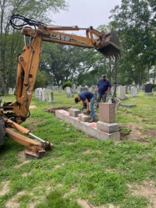 The bucket of an excavator hangs in the air as two men move headstones in the background